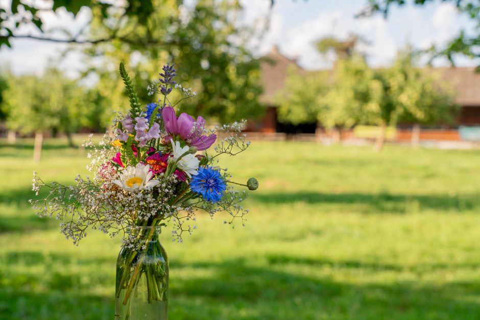 A glass jar filled with a hand-tied bouquet of fresh flowers, including pink tulips, white daisies, blue cornflowers, purple lupines, and delicate baby's breath, arranged with green stems and leaves. 
