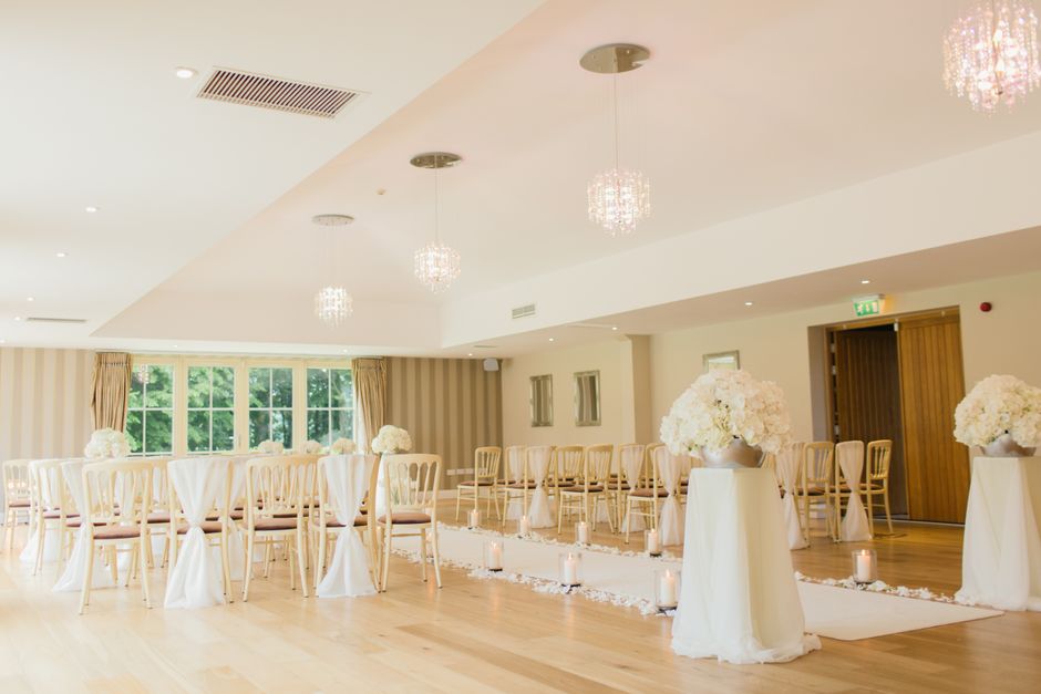 A spacious, well-lit wedding ceremony room decorated with floral arrangements. Several large white floral centerpieces consisting of hydrangeas and roses are displayed in white vases on tall, round ta