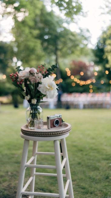 A tall, clear glass vase filled with a fresh floral bouquet featuring large white peonies, delicate pink and white roses, and clusters of small pink and white blossoms, complemented by lush green foli