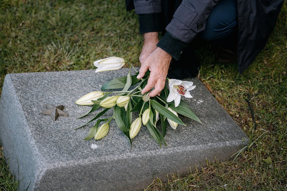 A person's hand placing a floral arrangement of white lilies with elongated petals and green leaves on a gray granite memorial stone, which features a small metal star emblem. The arrangement includes