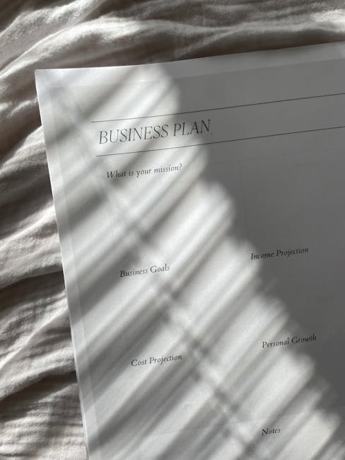 The image features a printed business plan document placed on a light-colored fabric surface, possibly a bed or tablecloth. The document is titled 'BUSINESS PLAN' and includes sections such as 'What i