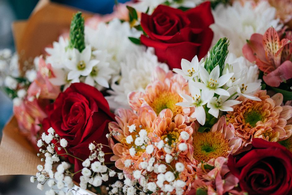 A vibrant floral bouquet featuring deep red roses, white star-shaped flowers, peach-colored chrysanthemums, and delicate white baby's breath, all arranged together with lush green foliage. The flowers