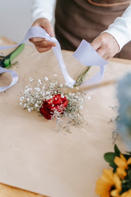 A person is arranging a floral gift on a light wooden table, holding a bouquet with a single vibrant red rose surrounded by delicate white baby's breath flowers. The bouquet is partially wrapped with 